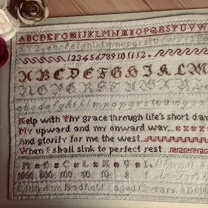 May include: A framed sampler with embroidered alphabets, numbers, and a verse in red and brown thread. The sampler includes the text "Help with Thy grace through life's short day." The sampler is dated 1080.