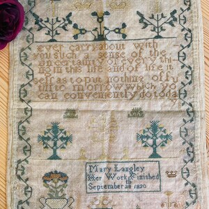 May include: Antique sampler with cross-stitch embroidery. The textile features a central inscription and decorative borders, with floral and tree motifs in green, yellow, and brown hues. The sampler is dated September 28, 1820, and includes the name Mary Langley.