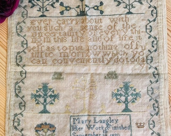 Könnte beinhalten: Antike Kreuzstich-Stickerei. Der Stoff zeigt eine zentrale Inschrift und dekorative Bordüren mit floralen und Baummotiven in Grün-, Gelb- und Brauntönen. Die Stickerei ist datiert auf den 28. September 1820 und enthält den Namen Mary Langley.