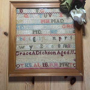 May include: A framed cross-stitch sampler in a wooden frame. The sampler displays rows of stitched text in colours like red, blue, and green. The text includes the name "Grace ADickson Aged //" and other words. Artificial flowers are attached to the frame.
