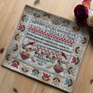May include: A vintage cross-stitch sampler with a cream background. The sampler features embroidered letters, numbers, and a verse in red and brown thread. Floral and bird motifs border the text, with the name "Angelena Morris" and the date "March 30, 1850" at the bottom.