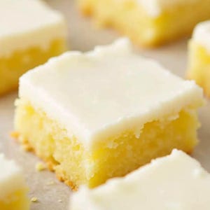 May include: Close-up of several square lemon bars with a thick, white, creamy frosting. The bars have a golden-yellow cake base and are arranged on a light-colored surface. The focus is on the dessert.