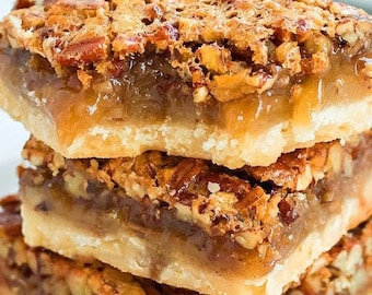 May include: A stack of three pecan pie bars with a flaky crust and a gooey pecan filling. The bars are topped with a layer of pecans and a drizzle of caramel.