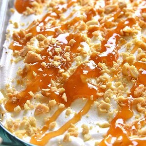 Op de afbeelding: Een dessert met lagen van graham cracker kruimels, slagroom en karamelsaus. Het dessert is bedekt met gehakte pinda's en meer karamelsaus.
