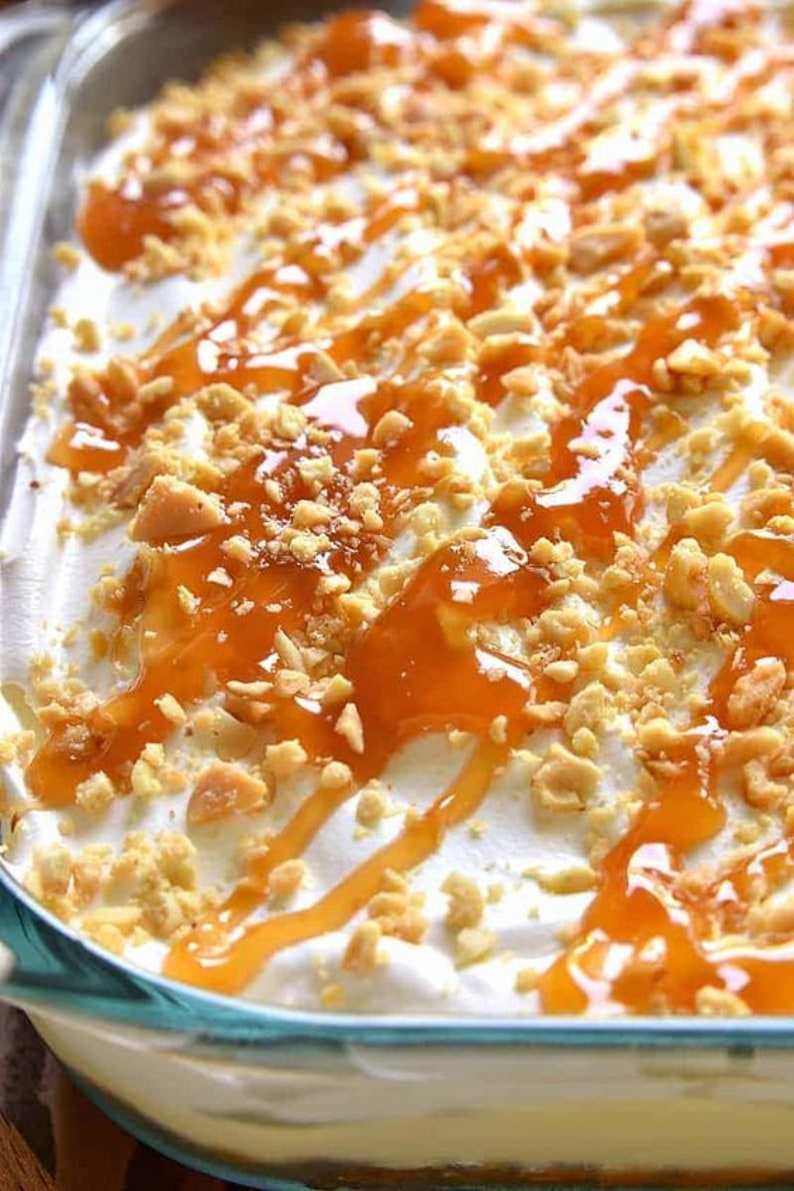 Op de afbeelding: Een dessert met lagen van graham cracker kruimels, slagroom en karamelsaus. Het dessert is bedekt met gehakte pinda's en meer karamelsaus.