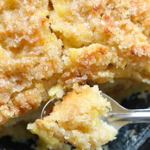 May include: Close-up of a golden brown baked dessert with a crispy, sugary topping. A spoonful of the dessert is being lifted out of a black baking dish.