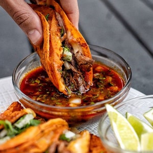 May include: A close-up of a hand dipping a crispy orange taco filled with meat and vegetables into a bowl of red sauce. The taco is being dipped into a bowl of red sauce. There are other tacos and lime wedges in the background.