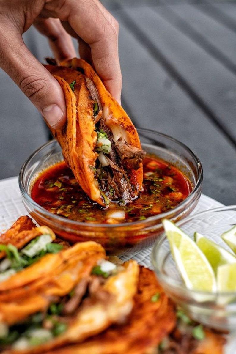 May include: A close-up of a hand dipping a crispy orange taco filled with meat and vegetables into a bowl of red sauce. The taco is being dipped into a bowl of red sauce. There are other tacos and lime wedges in the background.