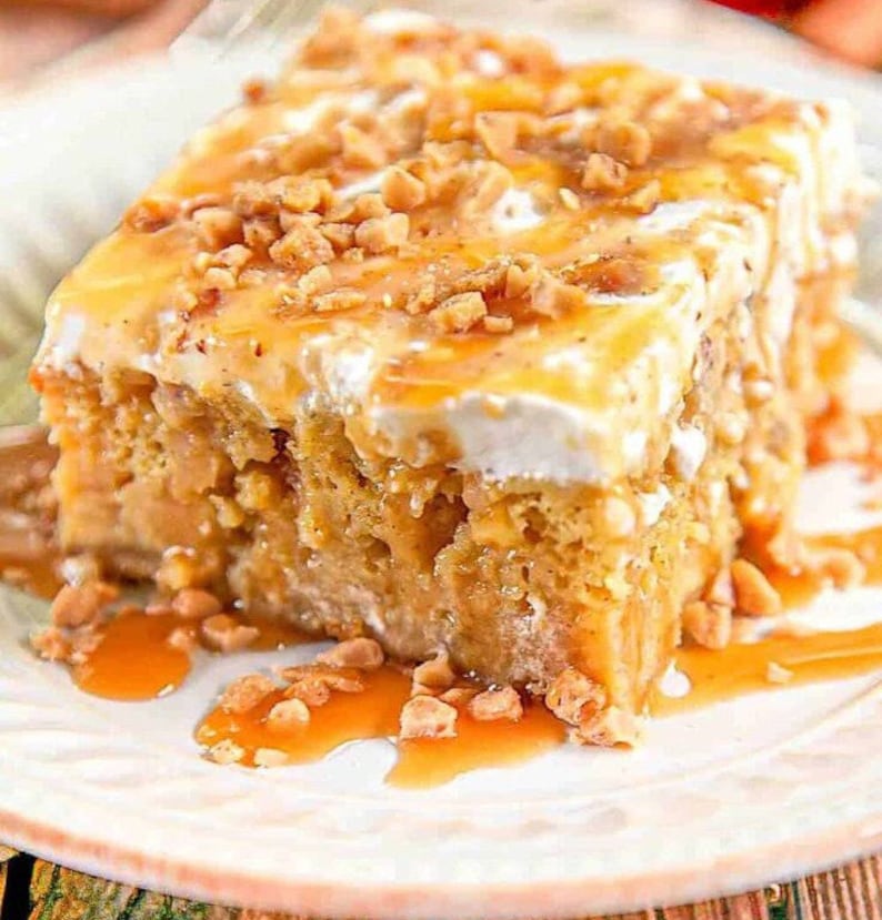 Op de afbeelding: Een plakje karamel appeltaart bedekt met slagroom en toffee stukjes. De taart ligt op een wit bord met een bruine achtergrond.