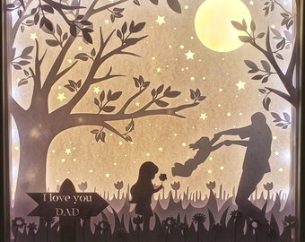 Op de afbeelding: Een kunstwerk van een silhouet dat uit papier is geknipt en een vader en dochter in een nachtelijke hemel voorstelt. De vader houdt de hand van zijn dochter vast terwijl ze in de lucht zwaait. De scène wordt verlicht door een heldere maan en sterren. Een bordje staat er: "Ik hou van je PAPA".