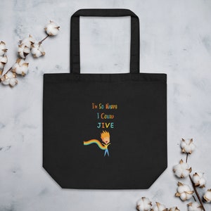 Puede incluir: Bolso tote negro con el texto "I'm So Happy I Could Jive" en letras coloridas. También aparece una figura de dibujos animados con una capa de arcoíris. El bolso tiene dos asas y está hecho de un material resistente.