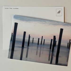 May include: A rectangular postcard featuring a photograph of dark wooden posts standing in calm, misty water at twilight. The sky is a soft gradient of pale pink and blue. The back of the card is white with the text Noordzee / Petten / Noord-Holland.