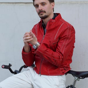 Könnte beinhalten: Rote Lederjacke mit weißen Nähten und Reißverschluss. Die Jacke wird mit einer weißen Hose und einer silbernen Uhr getragen. Im Vordergrund befindet sich ein schwarzes Fahrrad.