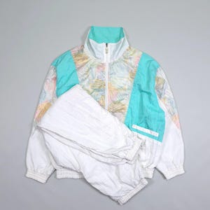 Può includere: Una tuta sportiva composta da una giacca a vento bianca e pantaloni bianchi abbinati. La giacca presenta un colletto alto, una chiusura frontale con zip integrale e pannelli color verde acqua sul petto e sulle maniche con un motivo pastello.