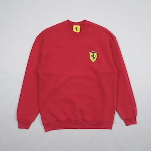 Może przedstawiać: Czerwona bluza z okrągłym dekoltem z logo Ferrari na piersi. Logo to czarny koń na żółtym tle. Bluza ma długie rękawy i prążkowane mankiety.