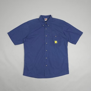 May include: A short-sleeved button-down shirt in a solid dark blue colour. The shirt features a button-down collar, a single chest pocket on the left side, and a small square patch on the pocket displaying a yellow graphic of a crescent moon.