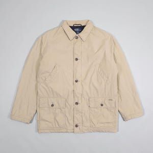 May include: A tan jacket with a button-up front, two chest pockets, and two lower pockets. The jacket has a collared neckline and long sleeves. The jacket is a neutral color and appears to be made of a lightweight material.