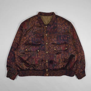 Puede incluir: Chaqueta bomber vintage con un diseño estampado en tonos marrón, burdeos y morado. La chaqueta presenta un cuello, botones dorados y dos bolsillos delanteros. Los puños y la cintura son acanalados.