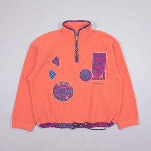 Könnte beinhalten: Korallenfarbener Fleece-Pullover mit hohem Kragen und Viertelreißverschluss. Der Pullover hat geometrische Aufnäher in Blau, Lila und Türkis. Der Saum und der Kragen sind mit einem gemusterten Stoff versehen. Der Text "FLORAL MAGIC" ist aufgedruckt.