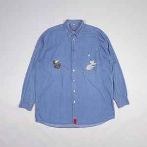 Puede incluir: Camisa vaquera azul claro de manga larga. Presenta un pequeño bolsillo en el pecho y bordados de animales de dibujos animados: un pájaro y un ratón. La camisa tiene un cuello clásico y una etiqueta roja en la parte inferior.