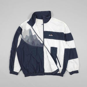 May include: A navy blue and white color-blocked windbreaker jacket featuring a high collar, a full-length front zipper, and two side pockets. The chest displays the ellesse brand logo, and the upper right panel includes a gray mountain graphic.