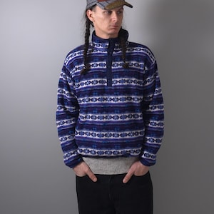 May include: A blue and white patterned fleece pullover with a high collar and a three-button placket. The pullover has a geometric design. The person is wearing a multi-colored baseball cap and black pants.