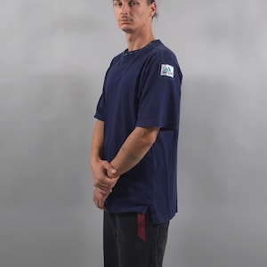 May include: A navy blue short-sleeved t-shirt featuring a small rectangular Adidas logo patch on the left sleeve. The shirt has a crew neckline with a subtle patterned trim visible at the collar and a straight hemline with side slits.