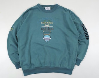 May include: A teal long-sleeve crewneck sweatshirt featuring embroidered text that reads Favorites Adidas Competition. The left sleeve displays the word Adidas in black lettering, while the right sleeve features smaller embroidered text.