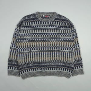 Può includere: Un maglione girocollo con un motivo geometrico nei toni del grigio, blu e marrone. Il maglione presenta un design ripetuto di linee verticali e orizzontali, creando un aspetto strutturato. Il colletto ha una piccola etichetta rossa.