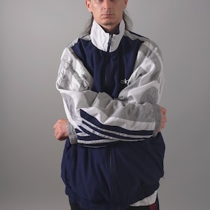 Puede incluir: Chaqueta deportiva vintage azul marino, blanca y gris con cierre de cremallera completo. La chaqueta presenta el logo de Adidas en el pecho y las mangas. Las mangas tienen rayas blancas y un cuello alto.