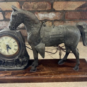 Puede incluir: Una figura de caballo de metal oscuro sobre una base de madera rectangular junto a un reloj circular. La esfera del reloj muestra números romanos y está enmarcada por una estructura metálica en forma de herradura. Una cadena une al caballo.