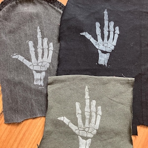 Könnte beinhalten: Drei Stoffflicken in Grau- und Schwarztönen, jeweils mit einer weißen Skeletthand mit erhobenem Mittelfinger. Die Flicken haben rohe, unfertige Kanten, was ihnen ein abgenutztes Aussehen verleiht. Das Design ist ein klares Statement.