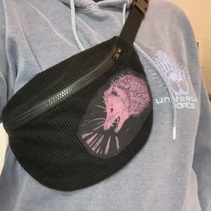Könnte beinhalten: Schwarze Cord-Bauchtasche mit Reißverschluss und verstellbarem Riemen. Auf der Vorderseite befindet sich ein Aufnäher mit einem rosa Opossum-Design. Die Bauchtasche wird über einem grauen Kapuzenpullover getragen. Der Text "UNIVERSE FORCE" ist sichtbar.
