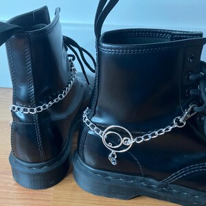 Könnte beinhalten: Paar schwarze Lederstiefel mit silbernen Ketten- und Charm-Akzenten. Die Stiefel haben ein klassisches Schnürdesign und eine robuste Sohle. Die silberne Kette umschließt den Knöchel und verleiht dem Schuhwerk einen Hauch von edgy Stil.