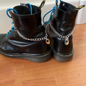 Könnte beinhalten: Paar schwarze Lederstiefel mit blauen Schnürsenkeln und silbernen Kettenakzenten. Die Stiefel haben eine glänzende Oberfläche und stehen auf einem hellen Holzboden.