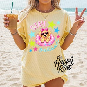 以下が含まれることがあります： 淡い黄色のTシャツ。ピンクのハート型サングラスとピンクのリボンをつけ、ピンクの浮き輪に入ったゴールデンレトリバーの子犬のグラフィックが描かれています。テキストは「Stay Golden」と「The Happy Riot」で、カラフルな星に囲まれています。