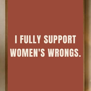 Könnte beinhalten: Ein rechteckiger Wandkunstdruck mit terrakottafarbenem Hintergrund und dem weißen Text "I FULLY SUPPORT WOMEN'S WRONGS." Der Druck ist in einem dünnen Holzrahmen eingefasst und hängt an einer Wand über einem hölzernen Konsolentisch.