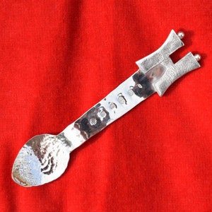 May include: A silver spoon with a hammered finish and a unique, abstract handle. The handle is shaped like a stylised letter H with a small ball at the top.