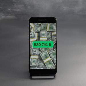 May include: A black smartphone displaying a screen filled with stacks of US dollar bills. The number "520 741 8" is displayed on the screen in a green rectangle.