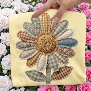 Puede incluir: Un cojín amarillo con un gran girasol aplicado. El girasol tiene un centro marrón con detalles bordados y pétalos de tela en varios patrones y colores, incluyendo azul, marrón y blanco. Las hojas son verdes. El cojín está sostenido por una mano.
