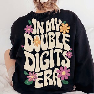 May include: Black sweatshirt with a white graphic that says "In My Double Digits Era" with colorful flowers around the text.