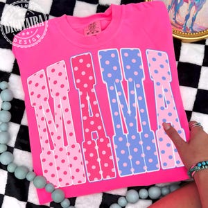 May include: A bright pink t-shirt with the word "MAMA" in large, polka-dotted letters. The letters alternate between pink and blue, with white outlines. The shirt has a crew neck and short sleeves.