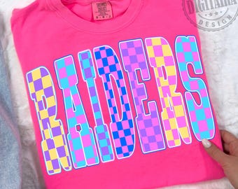 May include: A bright pink t-shirt with the word "RAIDERS" in large, checkered letters. The letters are filled with a pattern of blue, yellow, and purple squares. The shirt has a crew neck and short sleeves.