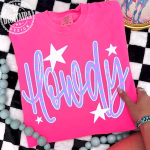 Puede incluir: Una camiseta rosa brillante con la palabra "Howdy" en un diseño a cuadros azul y morado, acentuada con estrellas blancas. La camiseta es de la marca Comfort Colors, y el diseño es de Digitalra Design.