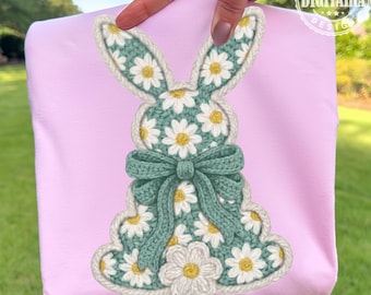 May include: A light pink fabric with a crochet bunny design. The bunny is decorated with white daisies and a green bow. The bunny's outline is white. The design is held by a hand.