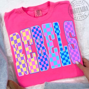 Può includere: Una maglietta rosa acceso con la scritta "REBELS" in grandi lettere a scacchi. Le lettere sono un mix di quadrati gialli, viola, blu e rosa. La maglietta è piegata e sembra essere fatta di materiale morbido.