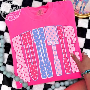 May include: A bright pink t-shirt with the word "AUNTIE" in large, polka-dotted letters. The letters alternate between pink and blue, each with a white outline. The shirt has a crew neck and short sleeves.