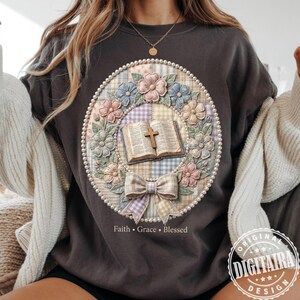 Könnte beinhalten: Dunkelgraues Sweatshirt mit floralem Kranzdesign. Der Kranz zeigt ein offenes Buch, ein Kreuz und pastellfarbene Blumen. Die Worte "Faith, Grace, Blessed" stehen darunter, umrahmt von einem perlenartigen Rand.