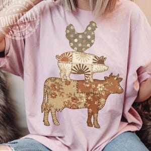 Könnte beinhalten: Hellrosa T-Shirt mit einem Patchwork-Design von Hoftieren. Eine braune Kuh mit floralen Akzenten trägt ein Schwein mit Sonnenstrahl-Details, gekrönt von einem gepunkteten Huhn. Das Design hat eine rustikale, ländliche Ästhetik.