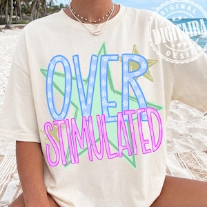 May include: Cream-colored t-shirt with the words "OVER STIMULATED" in a playful design. The word "OVER" is in light blue with white flower accents, and "STIMULATED" is in pink. The design is set against a green and yellow starburst.